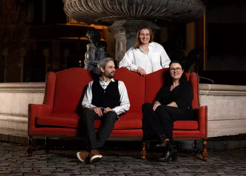 Drie mensen poseren op een rode bank voor een fontein in de avond.